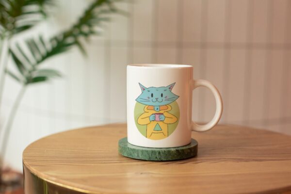 Cat Bud Mug is a plain white mug with an illustration of a cute blue cat sipping on a hot beverage